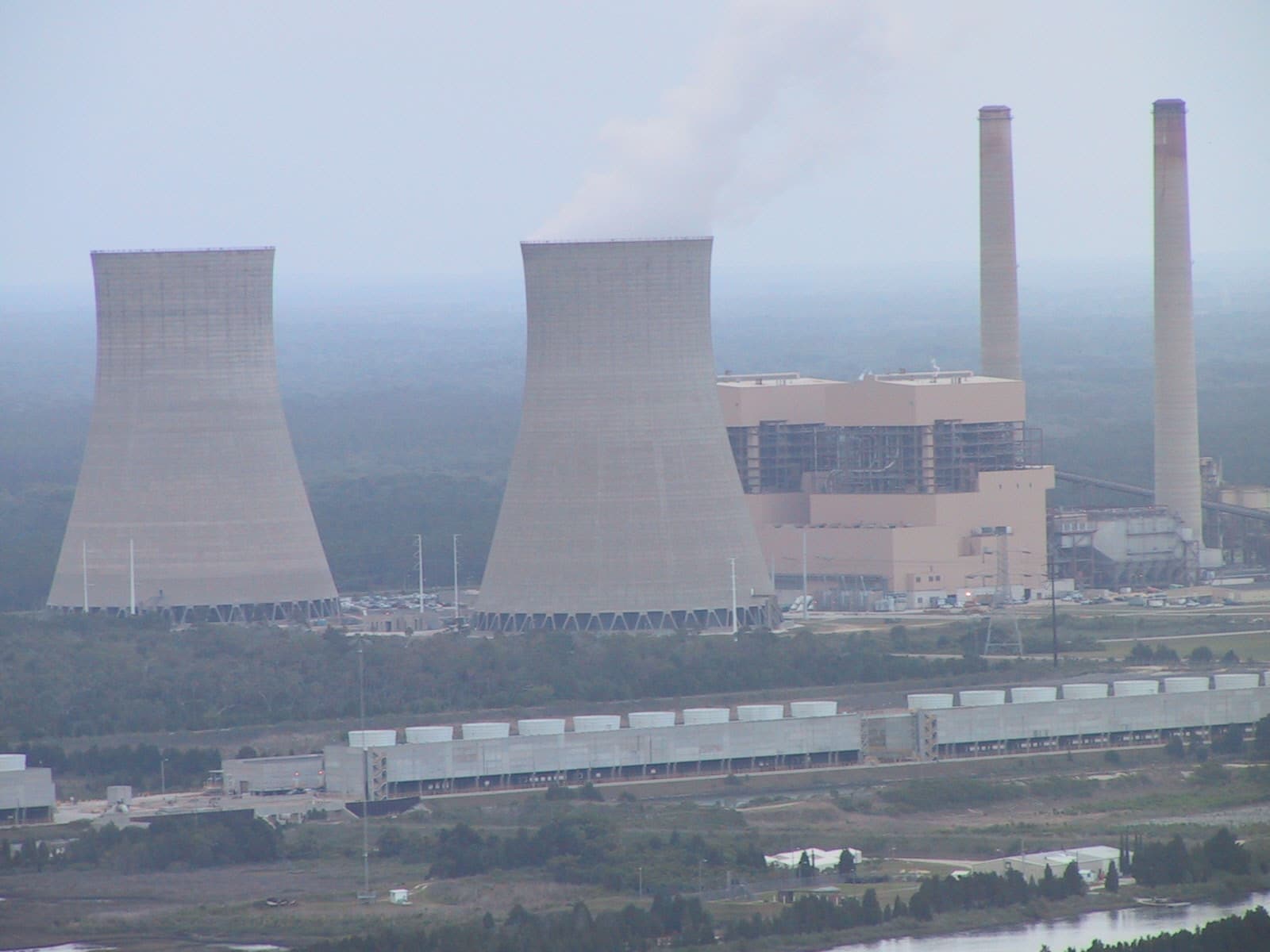 A power plant with two large cooling towers and two tall smokestacks surrounded by trees under a hazy sky