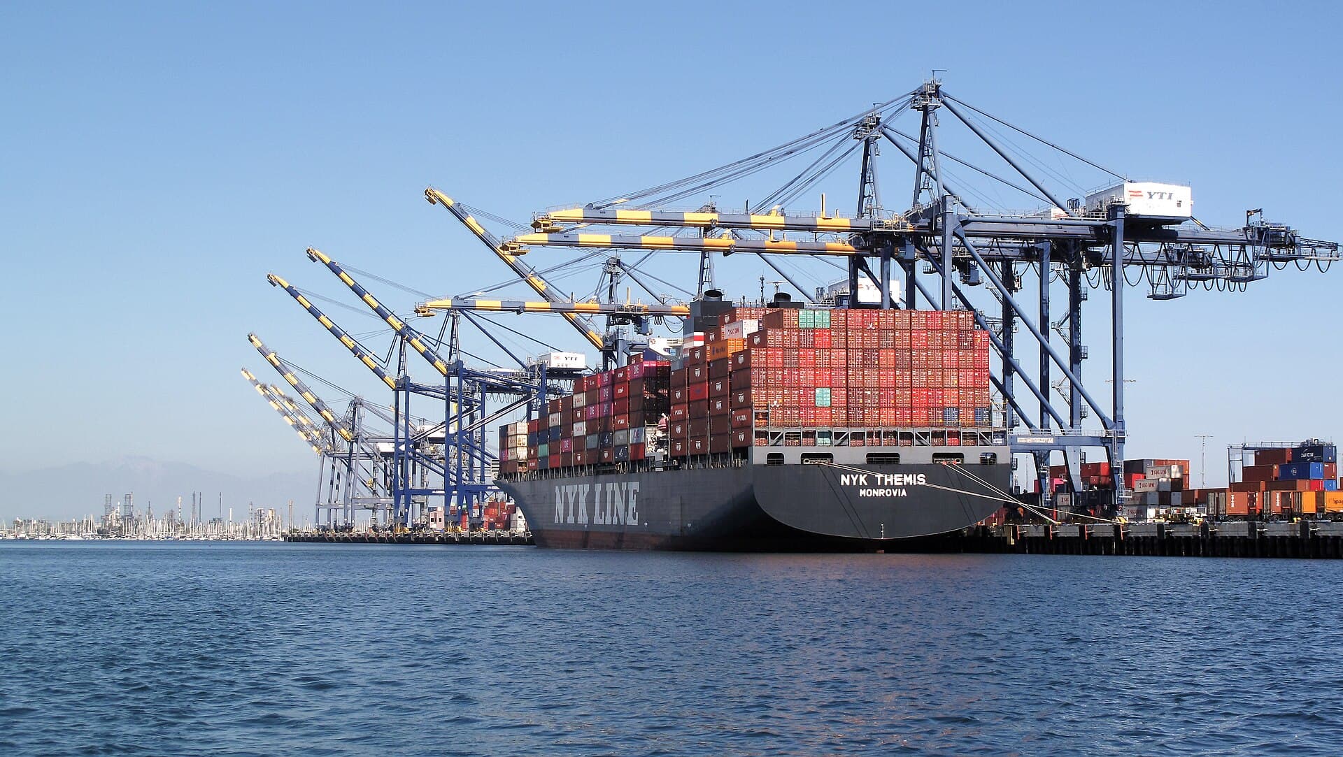 A container ship stacked with multicolored shipping containers beneath tall gantry cranes at a seaport