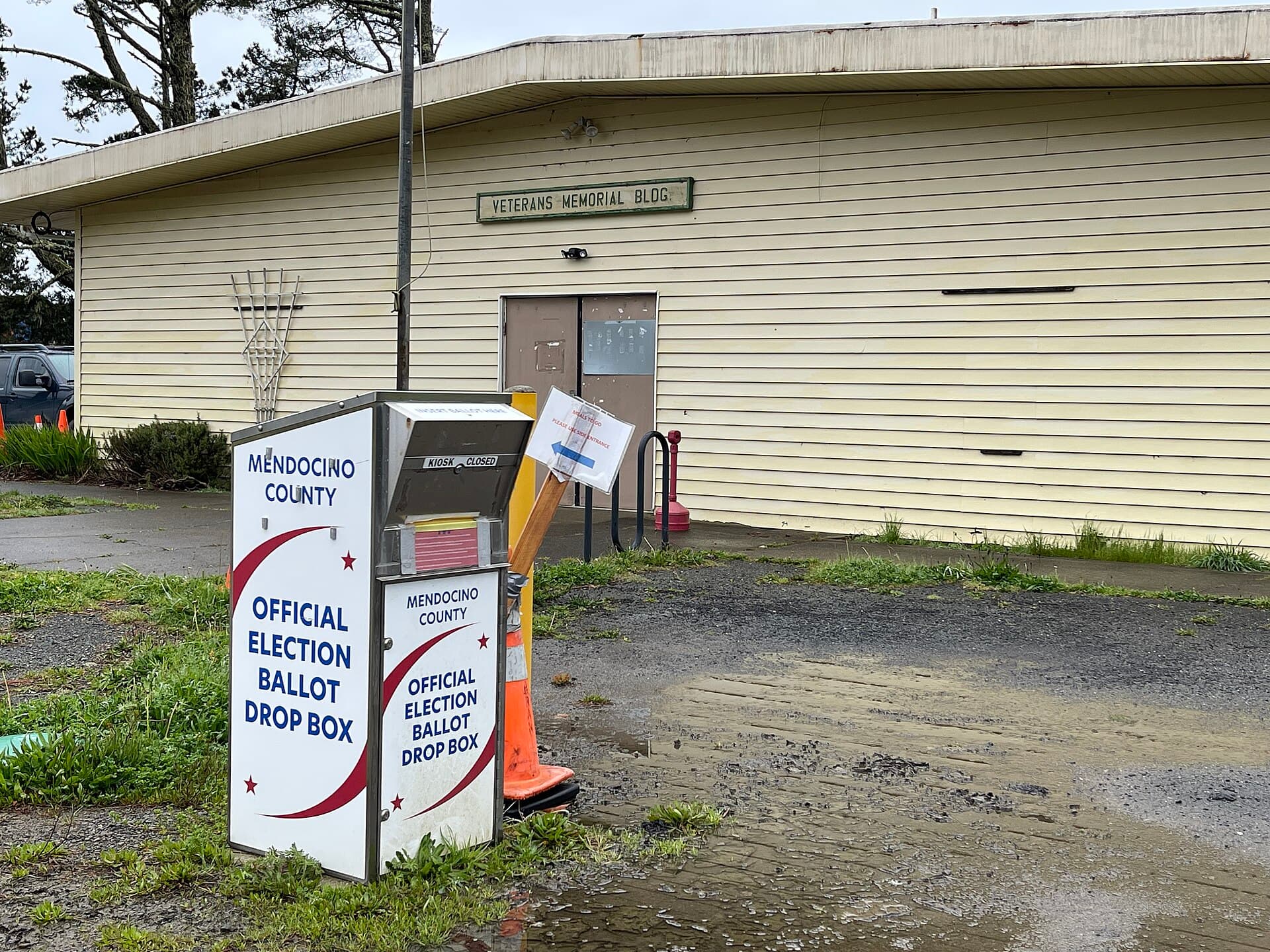 A metal official election ballot drop box standing outside a single-story civic building