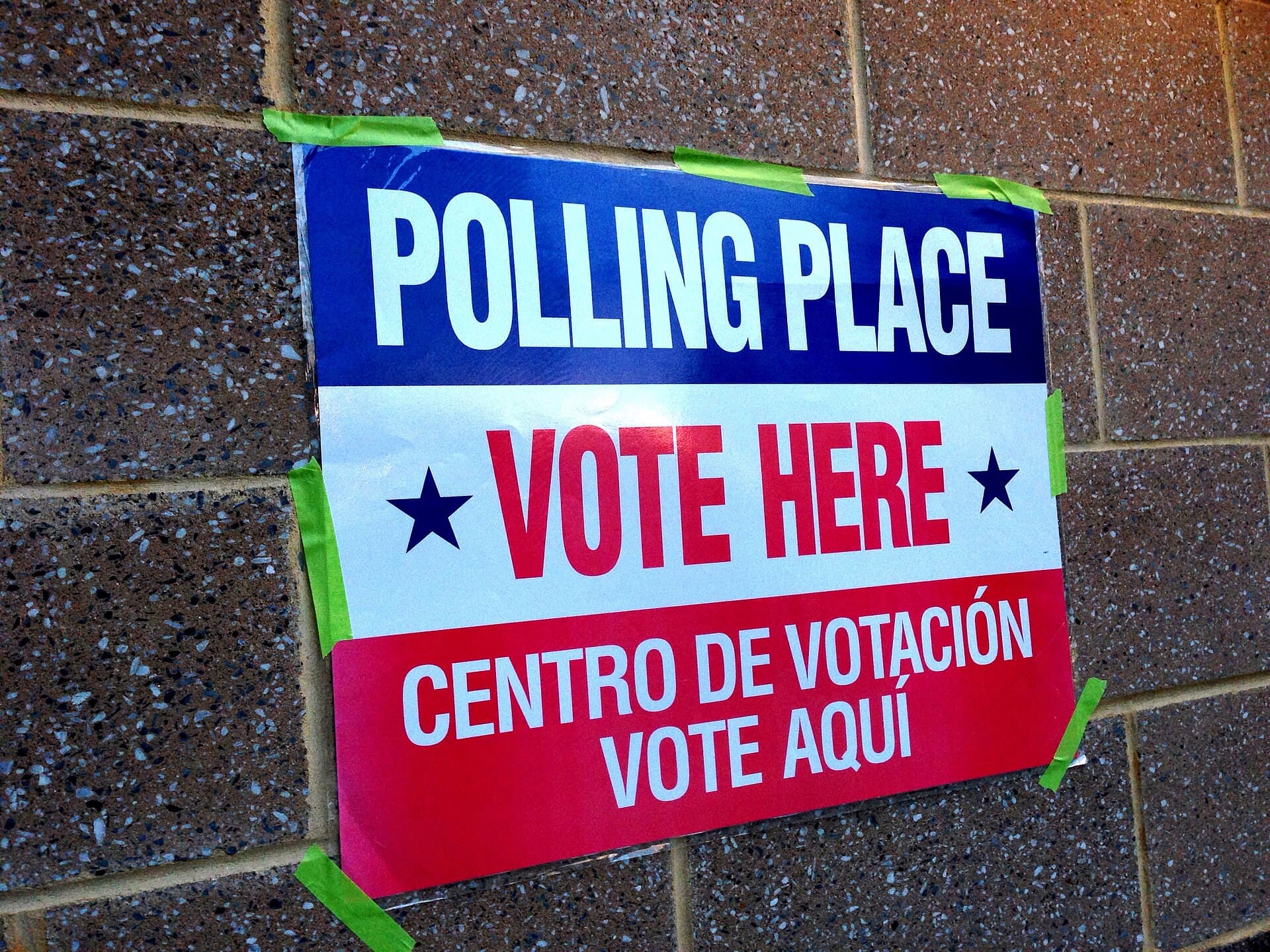 A red, white, and blue bilingual polling place sign reading Vote Here taped to a brick wall