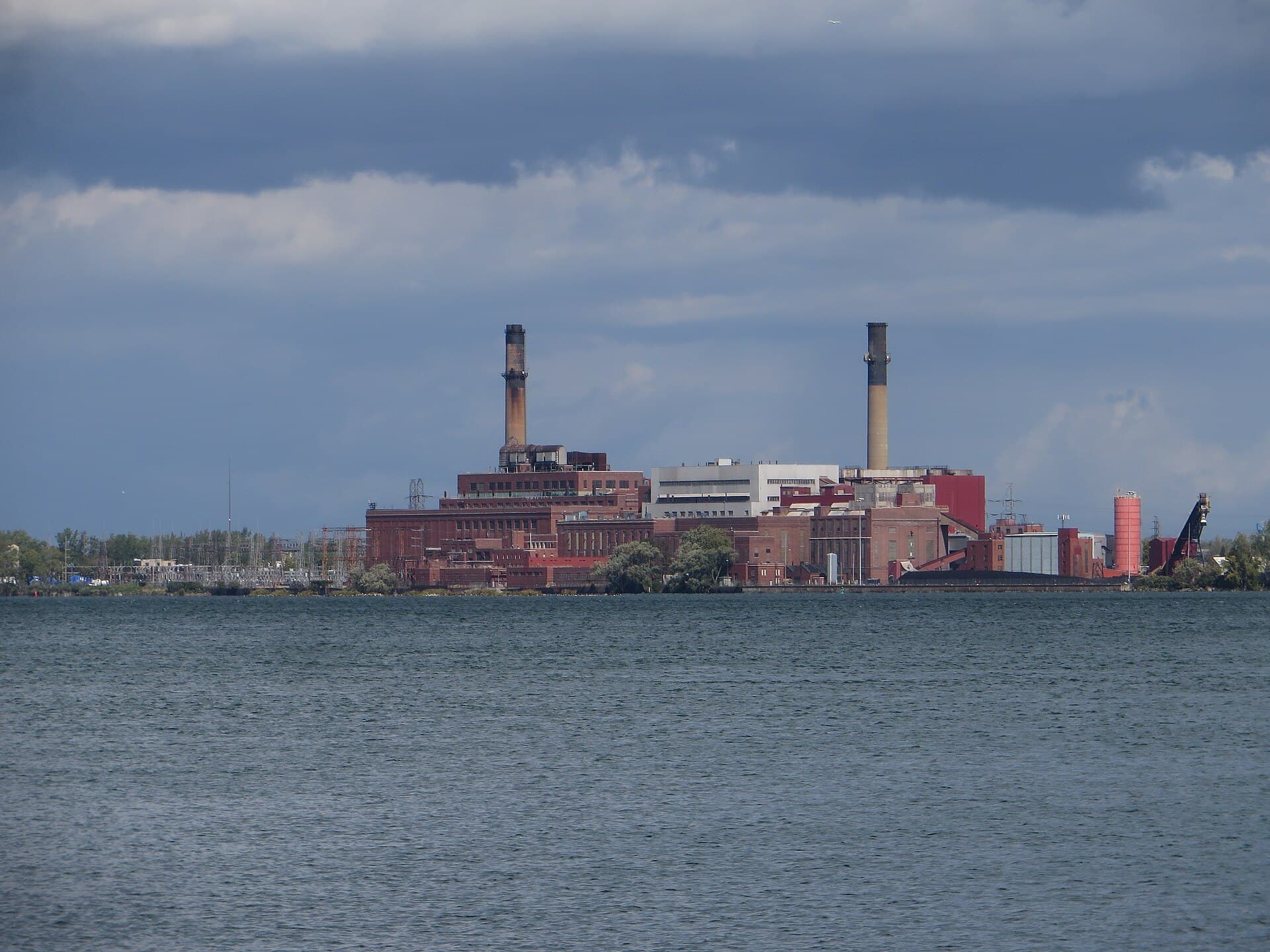 A brick power station with two tall smokestacks on a lakeshore under a cloudy sky