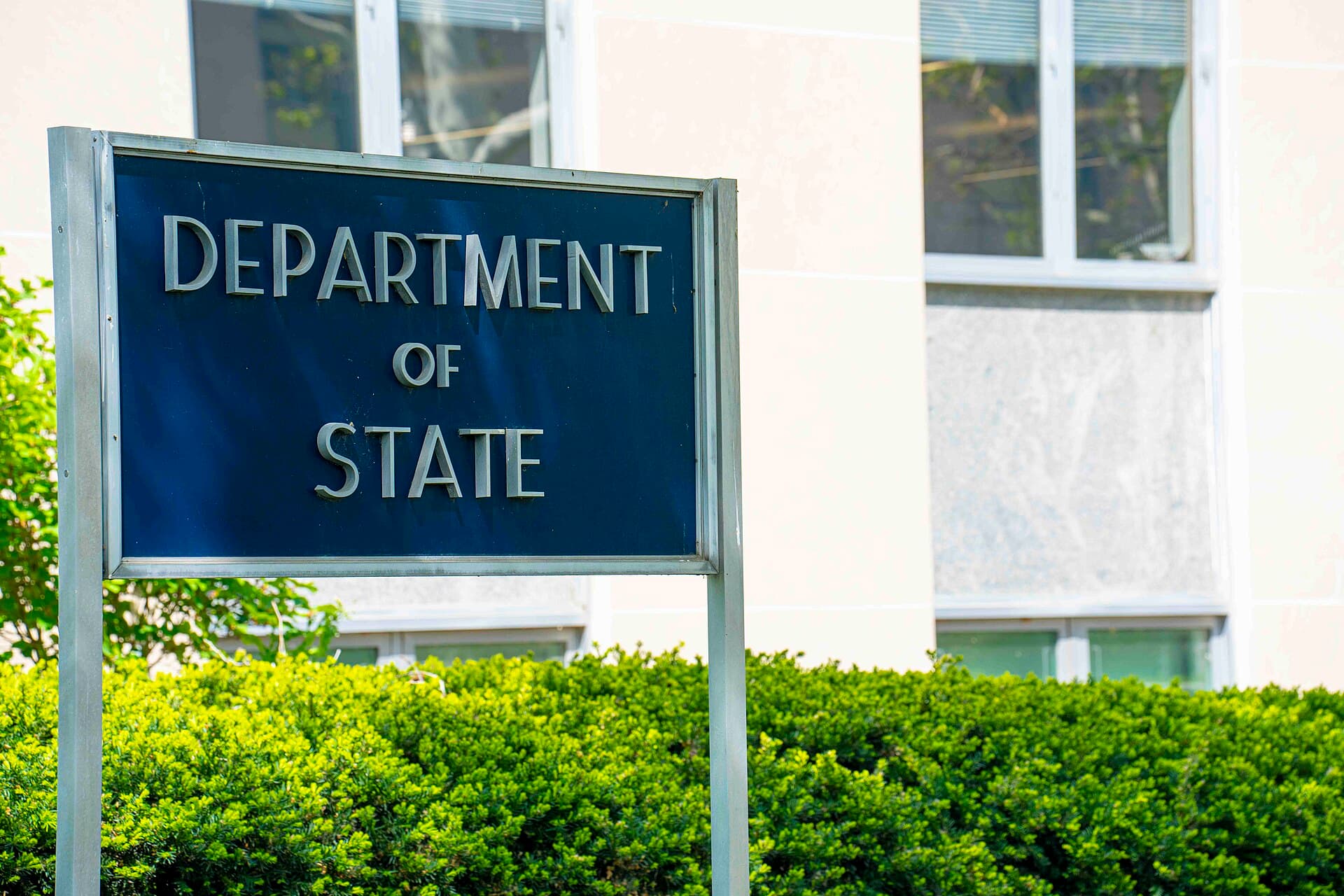 A dark blue Department of State sign in front of the agency's headquarters building with hedges below