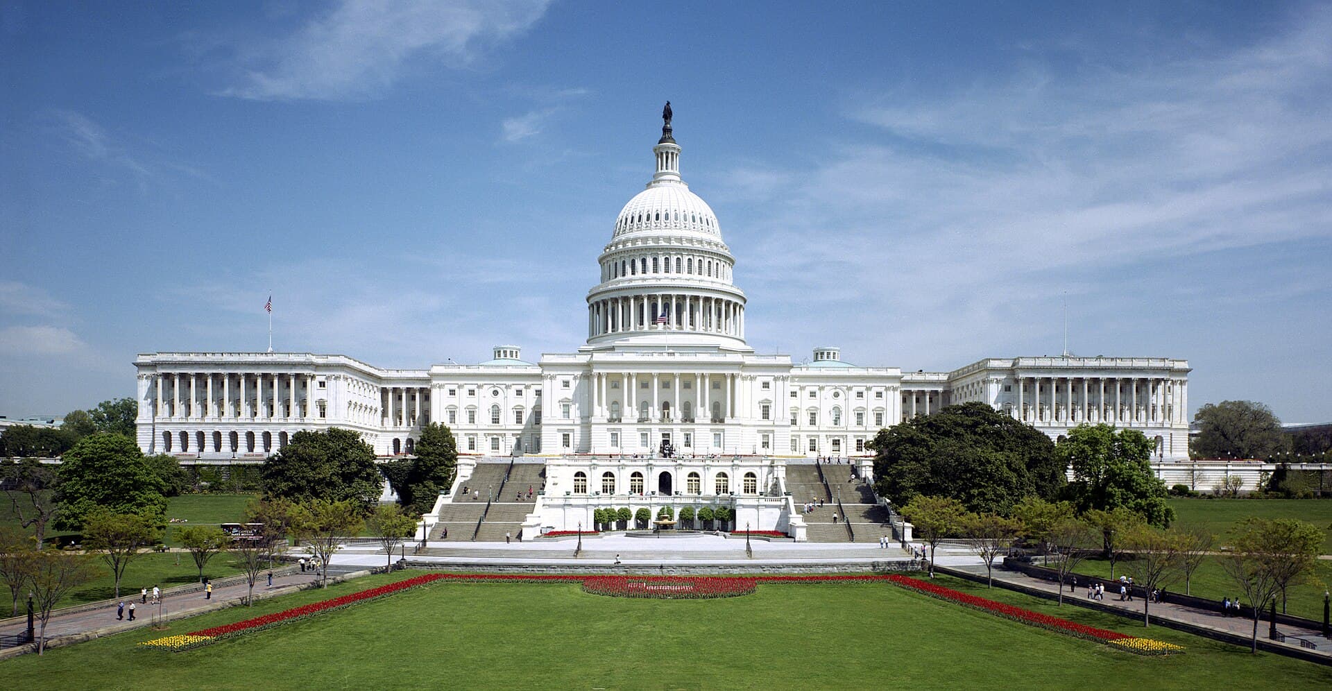 The west front of the United States Capitol with its dome, lawn, and flower beds under a blue sky